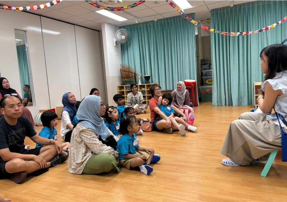 Adults and children sit on a classroom floor, listening to a woman seated on a chair at right.